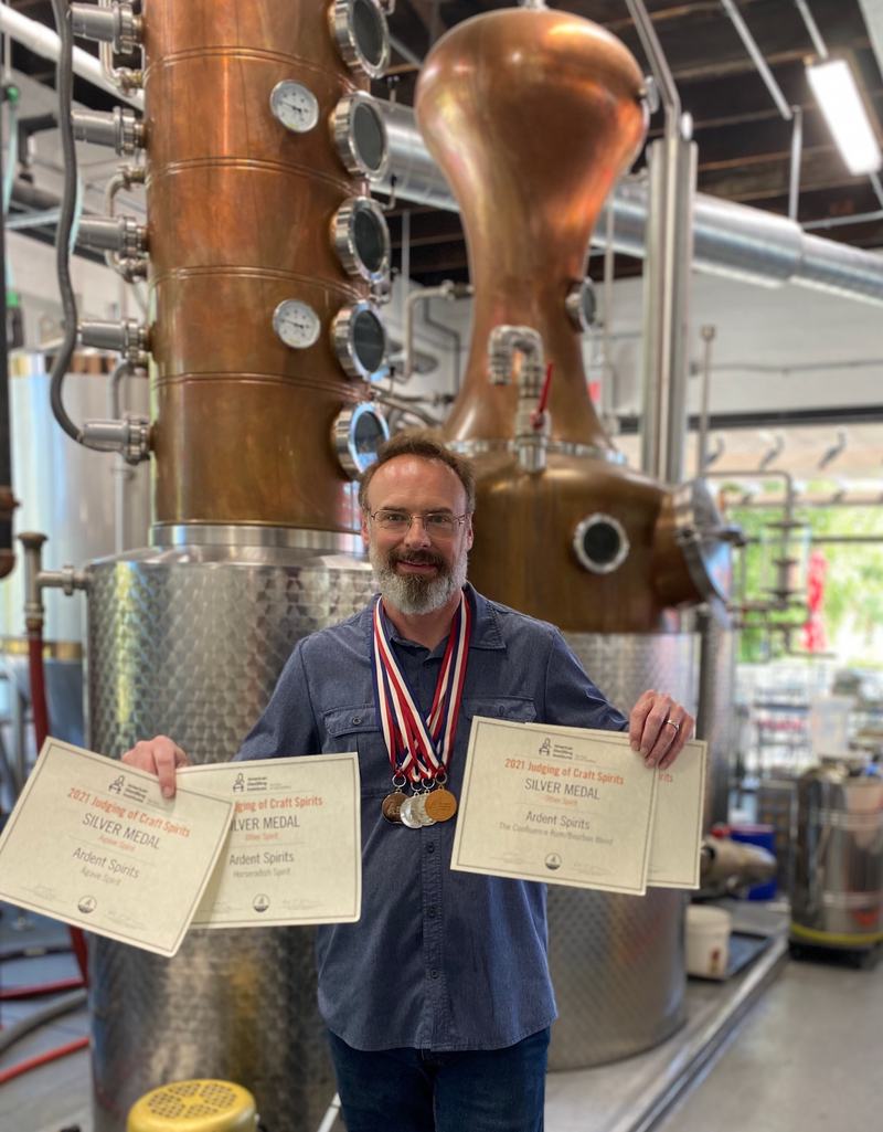 Derik, owner and distiller, standing in front of the copper still holding award certificates and wearing medals